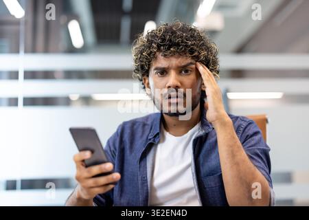 Close-up photo of a young Indian man holding a phone in his hand, standing in an office and looking worriedly into the camera. Stock Photo