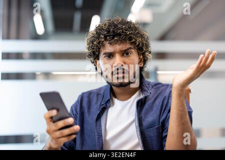 Close-up photo of a young Indian man holding a phone in his hand, looking at the camera with dissatisfaction and disappointment. Stock Photo