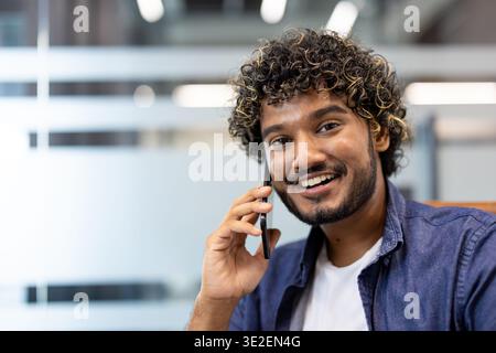 Close-up photo of a young Indian man sitting in an office and smiling while talking on the phone. Stock Photo