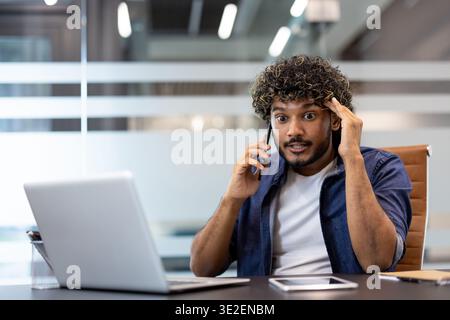 Worried young Indian man talking on the phone in the office, holding his head. Stock Photo