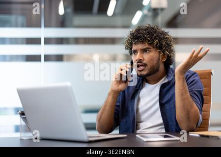 Angry and frustrated young Indian man sitting at a desk in the office and talking emotionally on a mobile phone. Stock Photo