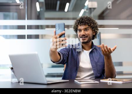 Young Indian man sitting at office desk and talking on video call via phone. Stock Photo