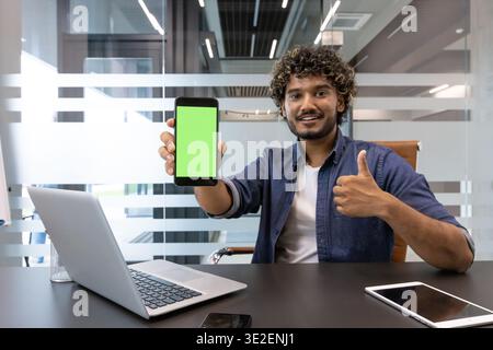 Portrait of an Indian young man in an office, sitting at a desk and showing a green screen mockup of a mobile phone to the camera. Stock Photo