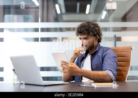 A serious young Indian man is sitting at a table with a laptop and looking thoughtfully at the tablet screen. Stock Photo