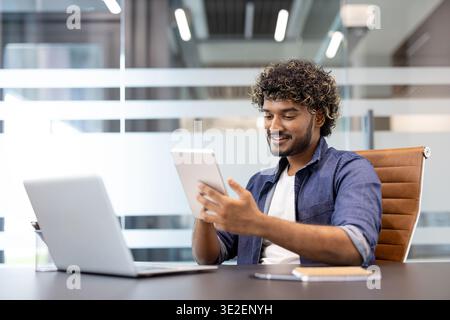A young Indian man is sitting in an office at a desk, working on a tablet. Stock Photo