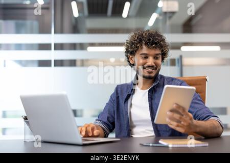 A young Indian man works in an office with a laptop and uses a tablet. Stock Photo