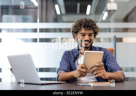 Smiling Indian man sitting at a desk with a laptop and working on a tablet in the office. Stock Photo