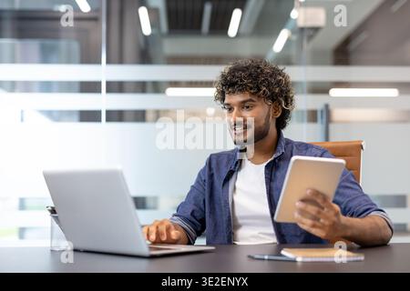 Smiling young Muslim man working in the office at a laptop and using a tablet. Stock Photo