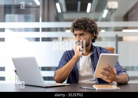 Thoughtful Indian male office worker working on tablet and laptop. Stock Photo