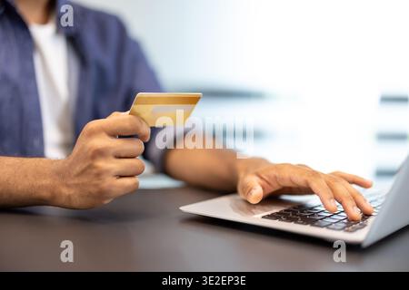 Close-up photo of the hands of a young man sitting at a desk in an office, holding a gold credit card and using a laptop. Stock Photo