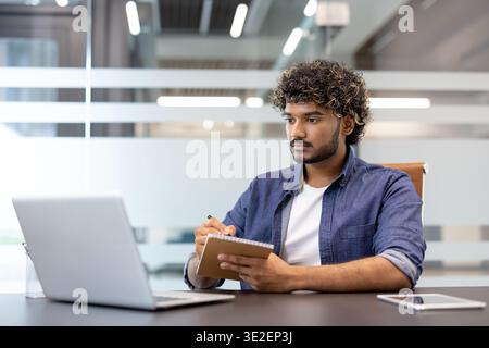 A focused young man is sitting at a desk in the office, looking at the laptop screen and making notes in a notebook. Stock Photo