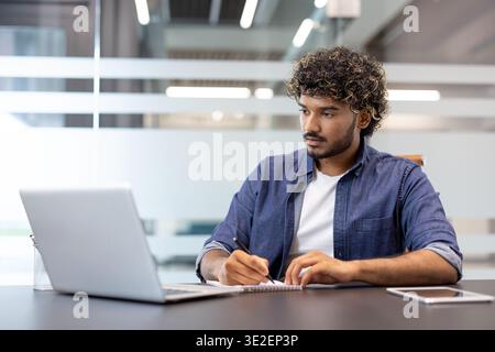 A young Indian man works in an office at a desk and takes notes in a notebook. Stock Photo