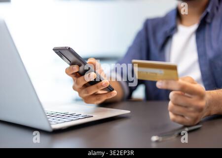 Close-up photo of the hands of a young man sitting at a desk with a laptop, holding a credit card in his hand and using a mobile phone. Stock Photo
