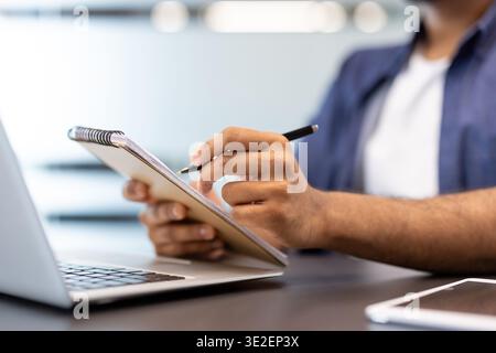 Close-up photo of the hands of a young man working in the office on a laptop and taking notes in a notebook. Stock Photo