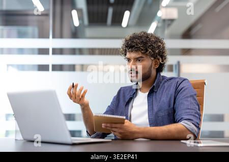 Worried young Indian man sitting at a desk in the office, holding a tablet in his hands and looking at the laptop screen in frustration. Stock Photo