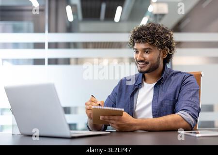 A young Indian man is sitting in an office at a desk, looking at a laptop screen and making notes in a notebook. Stock Photo