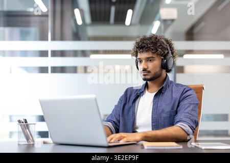 Concentrated young Indian man wearing headphones working on laptop in office. Stock Photo