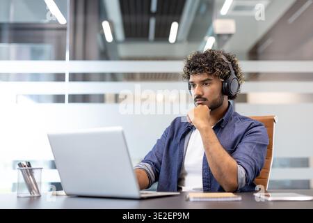 Serious and focused young Indian man in headphones working in the office on a laptop. Stock Photo
