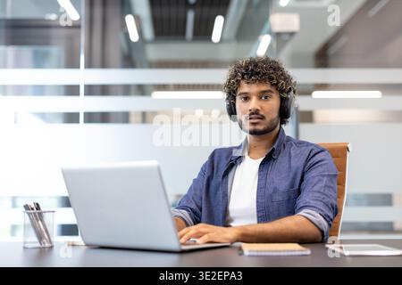 Portrait of a young Indian man wearing headphones working or studying on a laptop in the office, looking seriously at the camera. Stock Photo