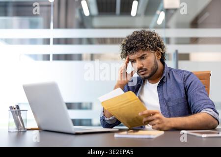 Upset and disappointed Indian young man sitting at the desk in the office and reading the letter he received. Stock Photo