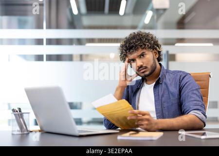 A sad young Indian man sits at a desk in the office, holds his head and anxiously reads the letter he received. Stock Photo