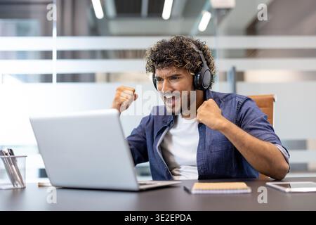Happy young Indian man sitting at the desk in the office wearing headphones, looking at the laptop screen, rejoicing and cheering. Stock Photo