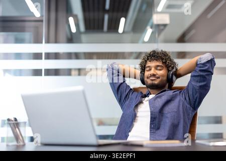 Satisfied young Indian man sitting at office desk wearing headphones with eyes closed and relaxing. Stock Photo