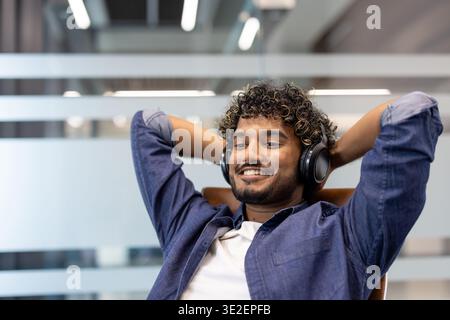 Relaxed young Indian man wearing headphones sitting in a chair in the office, hands behind his head, resting. Stock Photo