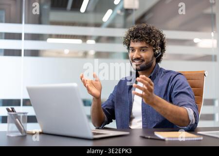 A young Muslim man is sitting at a desk in an office wearing a headset and talking on a video call on a laptop. Stock Photo