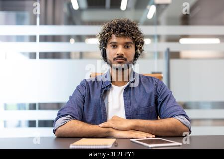 Photo of a young Indian man wearing headphones sitting at a desk in an office facing the camera and making a video call. Stock Photo