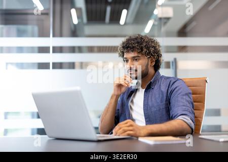 Serious young thoughtful Indian man working in the office on a laptop. Stock Photo
