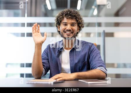 Close-up portrait of a young smiling Indian man sitting in an office at a desk facing the camera and raising his right hand. Stock Photo