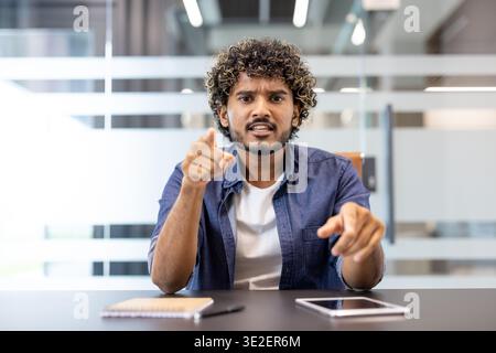 Portrait of a young angry Indian man sitting in the office facing the camera and talking emotionally. Stock Photo