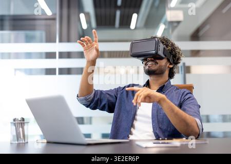 Young Indian man wearing virtual reality glasses sitting at a desk in an office and gesturing with his hands. Stock Photo