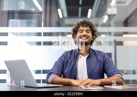 Portrait of a young Indian man sitting in the office at his desk facing the camera with his eyes closed and resting contentedly. Stock Photo