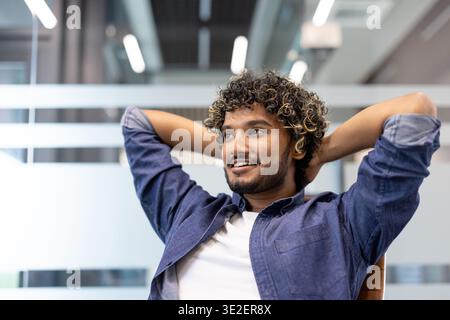 Close-up photo of a young Indian man sitting in a chair at an office desk with his hands behind his head, smiling. Stock Photo