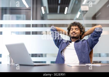 Relaxed young Indian man with his hands behind his head and eyes closed resting in a chair in the office. Stock Photo