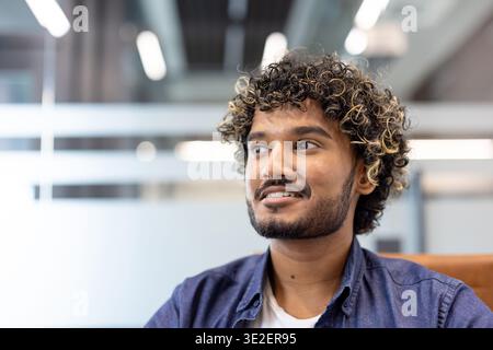 Close-up photo of a young Indian man sitting in an office and looking to the side with a smile. Stock Photo