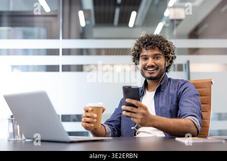 Portrait of a young smiling Indian man sitting in the office at a table with a laptop, holding a coffee in his hand and looking at the camera. Stock Photo