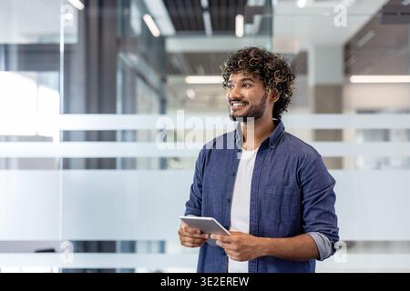 Smiling young Indian man standing in office, using tablet and looking to the side. Stock Photo