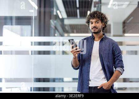 Portrait of a young Indian man standing in an office, holding his hand in his pocket, using his phone and looking at the camera. Stock Photo