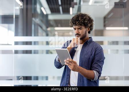 A thoughtful and serious Indian man is standing in an office and looking at the tablet screen. Stock Photo