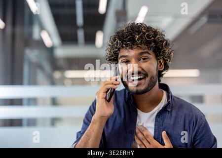 Close-up photo of an Indian young man in an office, looking at the camera and talking emotionally on the phone. Stock Photo