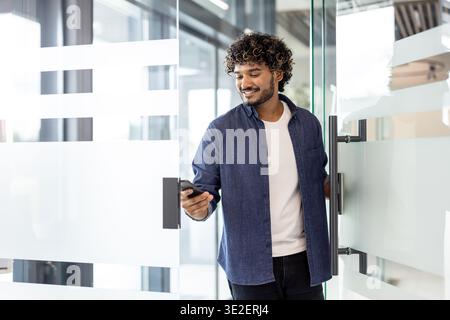 An Indian smiling man holds a phone in his hands, opens the door and enters the office premises. Stock Photo