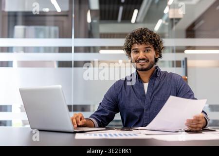 Portrait of a smiling and successful Indian businessman sitting in the office at a table with a laptop, holding documents in his hands and looking at the camera. Stock Photo