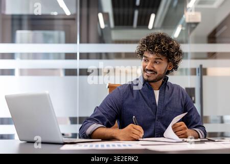 Smiling young Indian man working in the office with documents, looking at the laptop screen and making notes on paper. Stock Photo