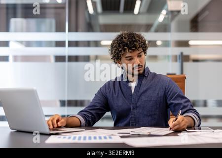 A serious young Indian man is working intently in the office on a laptop and making notes in documents. Stock Photo
