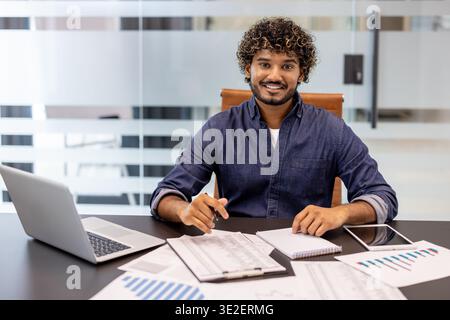 Portrait of a smiling young Indian man sitting at a desk in the office, working with documents and looking at the camera. Stock Photo