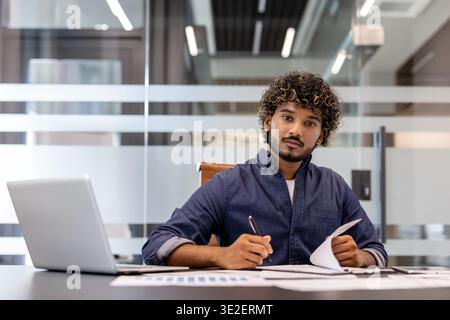 Portrait of a serious young Indian businessman working in the office with a laptop and documents, looking at the camera. Stock Photo