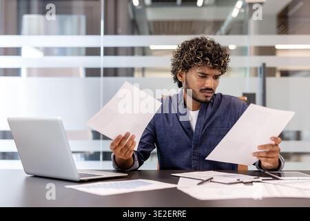 Serious young Indian man working in the office with a laptop and documents. Stock Photo